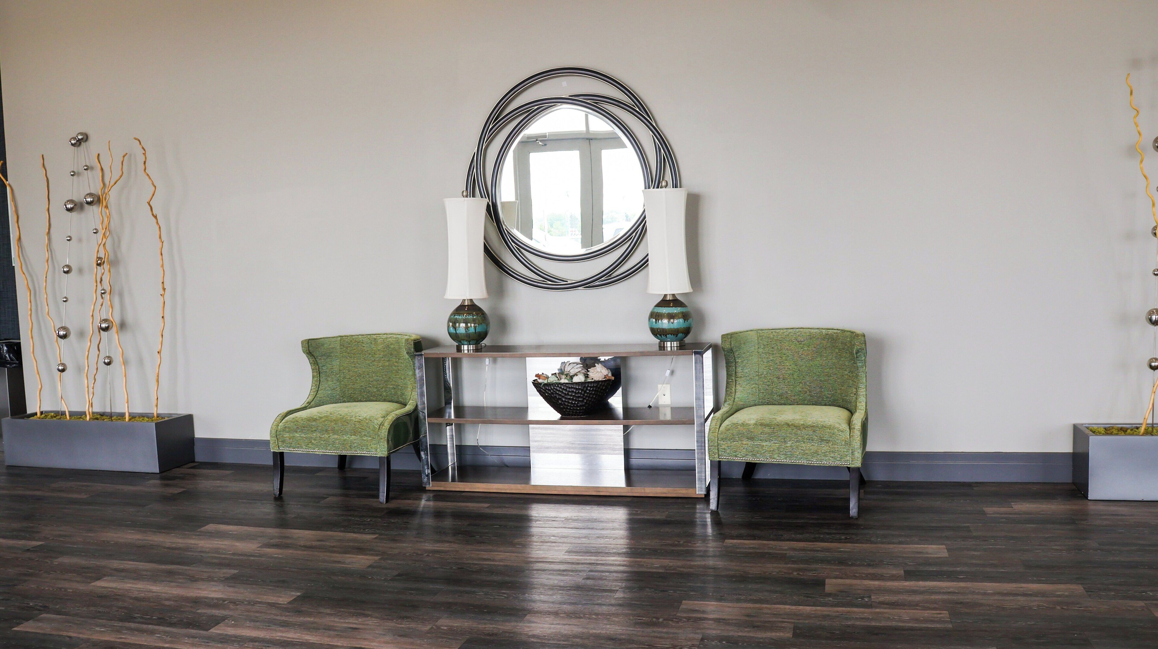 A modern living room with a dark wood floor, two green upholstered chairs, a glass console table with decorative items, a large mirror, and abstract wall art.