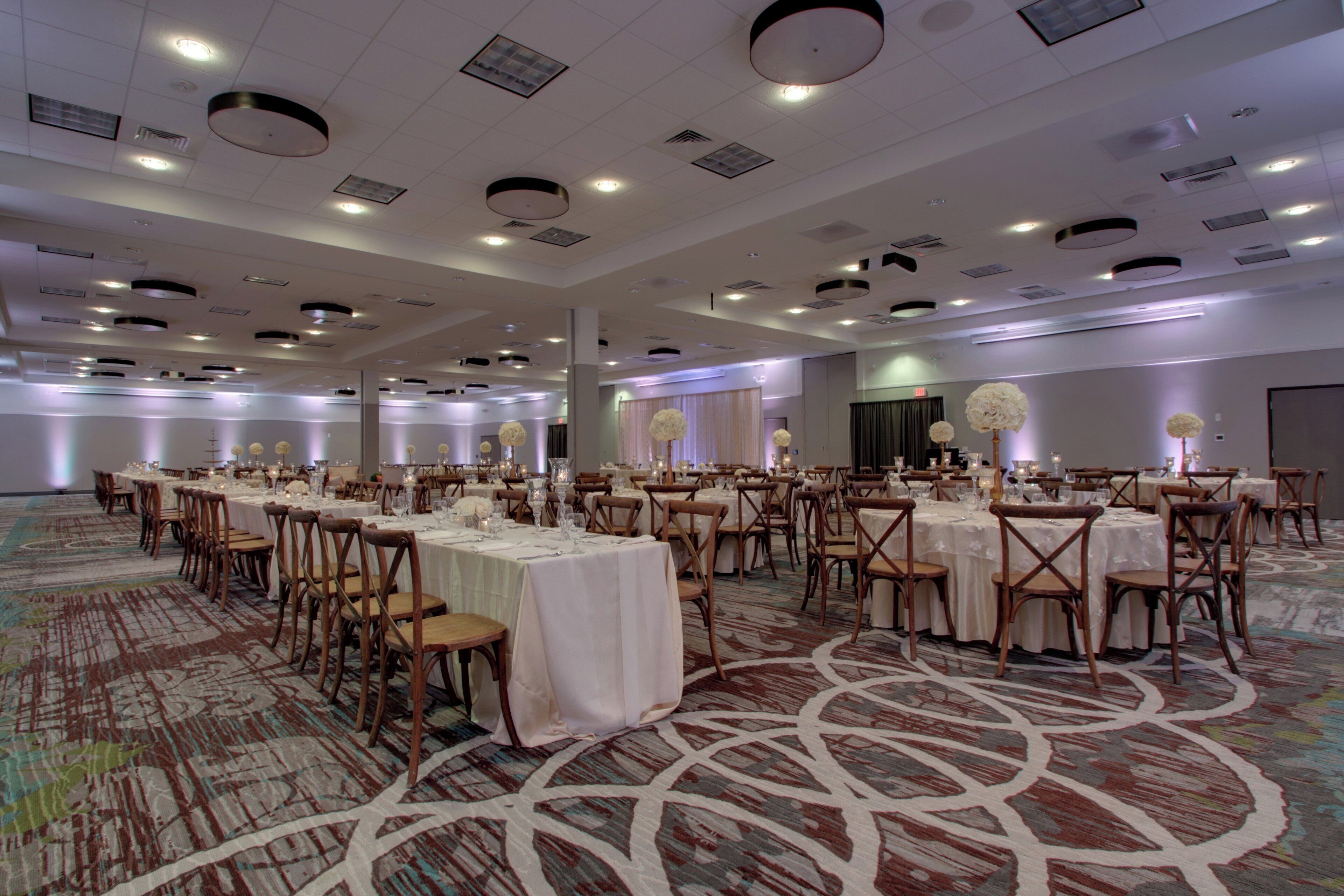 A large banquet hall with multiple round tables, each set with white tablecloths and chairs, and a patterned carpet.
