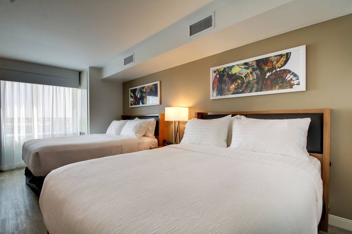 A hotel room with two beds, a large window with sheer curtains, and two abstract paintings on the wall.
