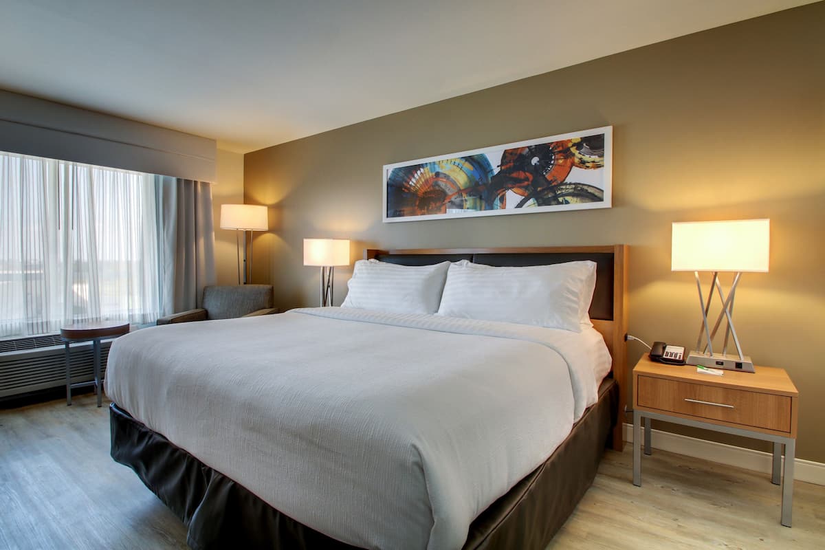 A hotel room with a large bed, two bedside tables with lamps, a window with curtains, and a painting on the wall.