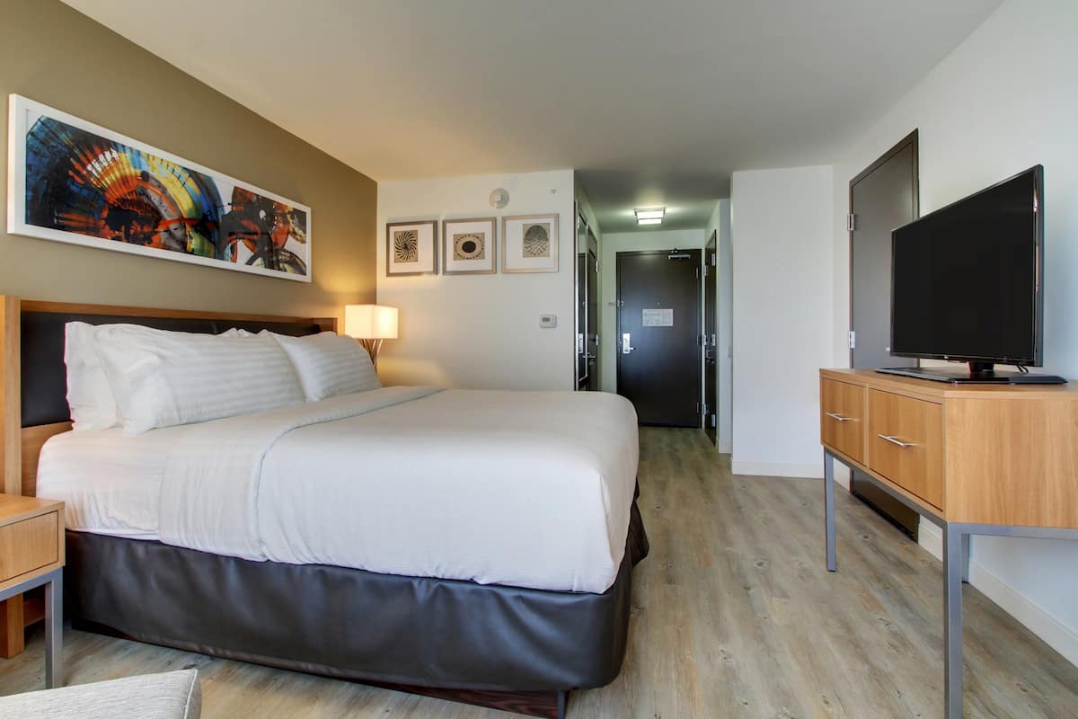 A hotel room with a bed, a TV, a desk, and a painting on the wall.