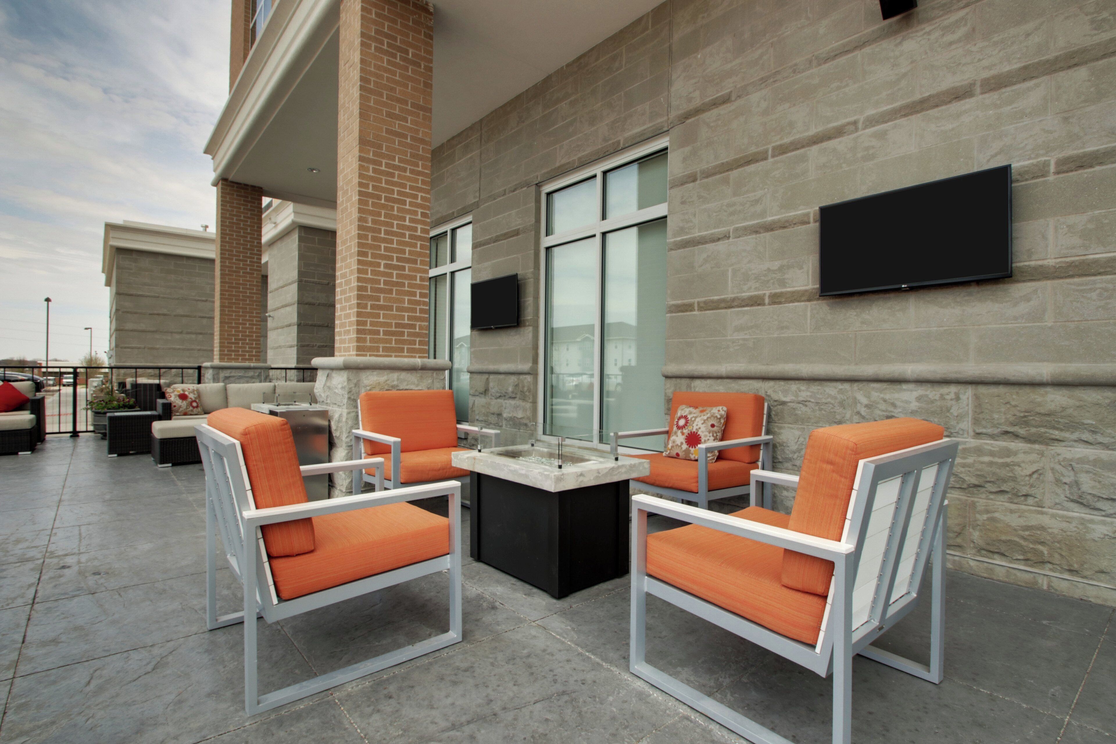 An outdoor seating area with two chairs and a table, a flat-screen TV mounted on the wall, and a brick wall in the background.