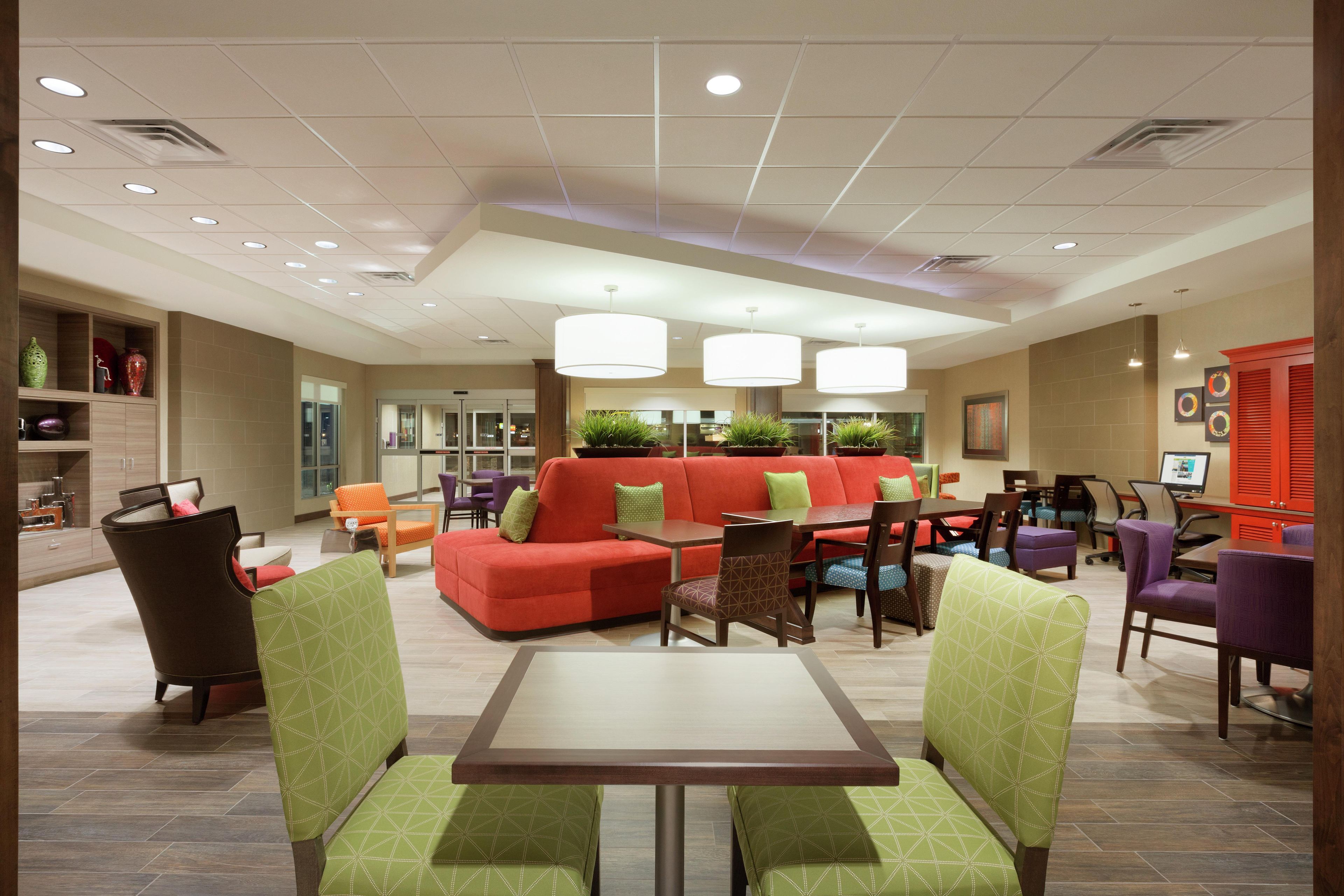 A modern hotel lobby with a red sofa, green chairs, a dining table, and pendant lighting.