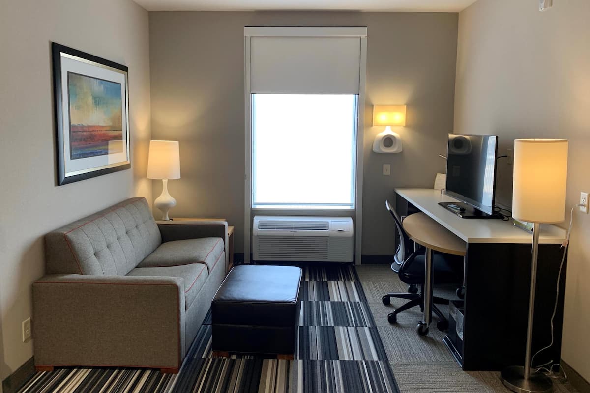 A hotel room with a sofa, a desk with a TV, a lamp, a picture on the wall, and a window with blinds.