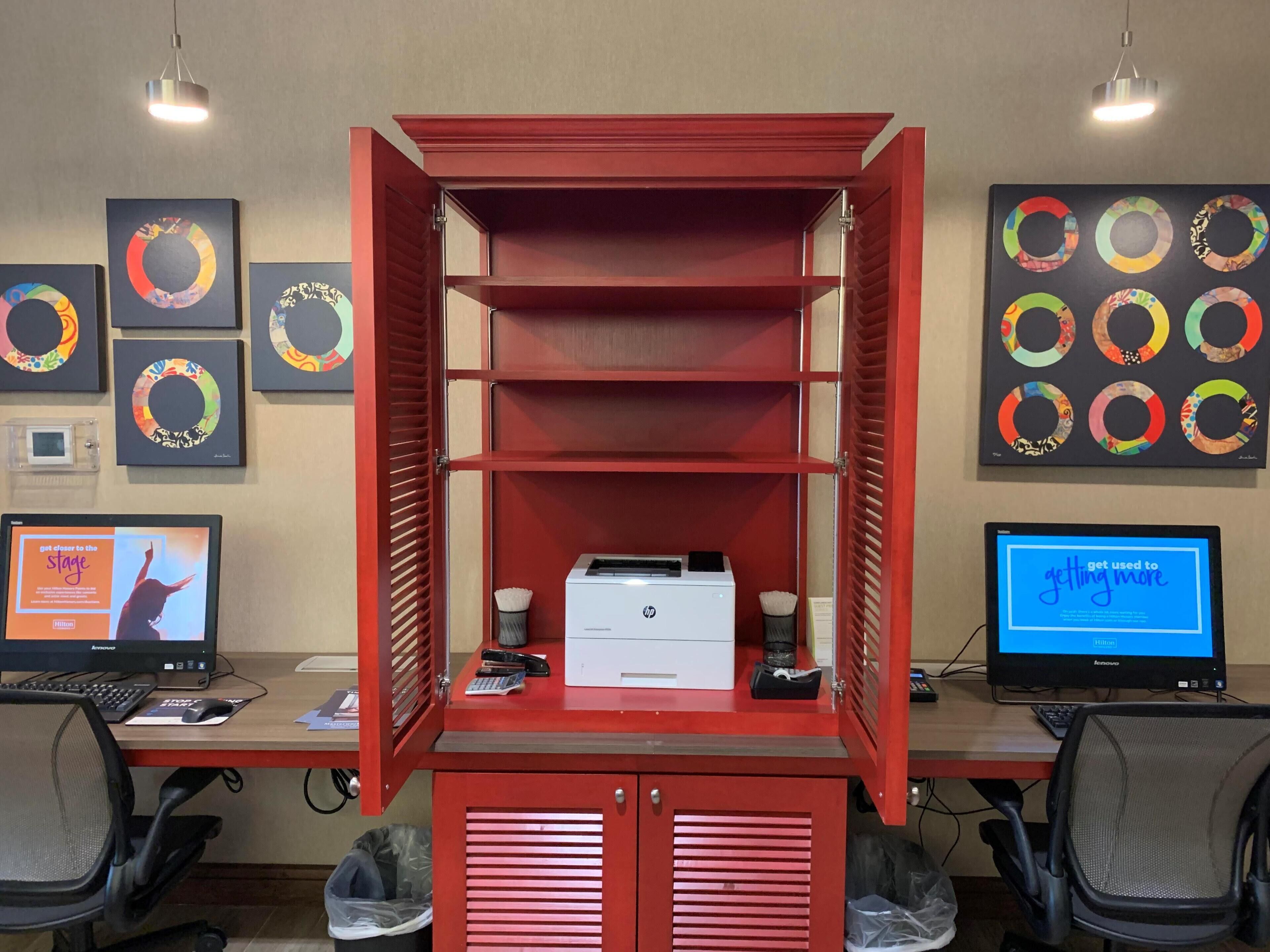 A hotel room with a red cabinet, two monitors, a keyboard, a mouse, and a trash bin.
