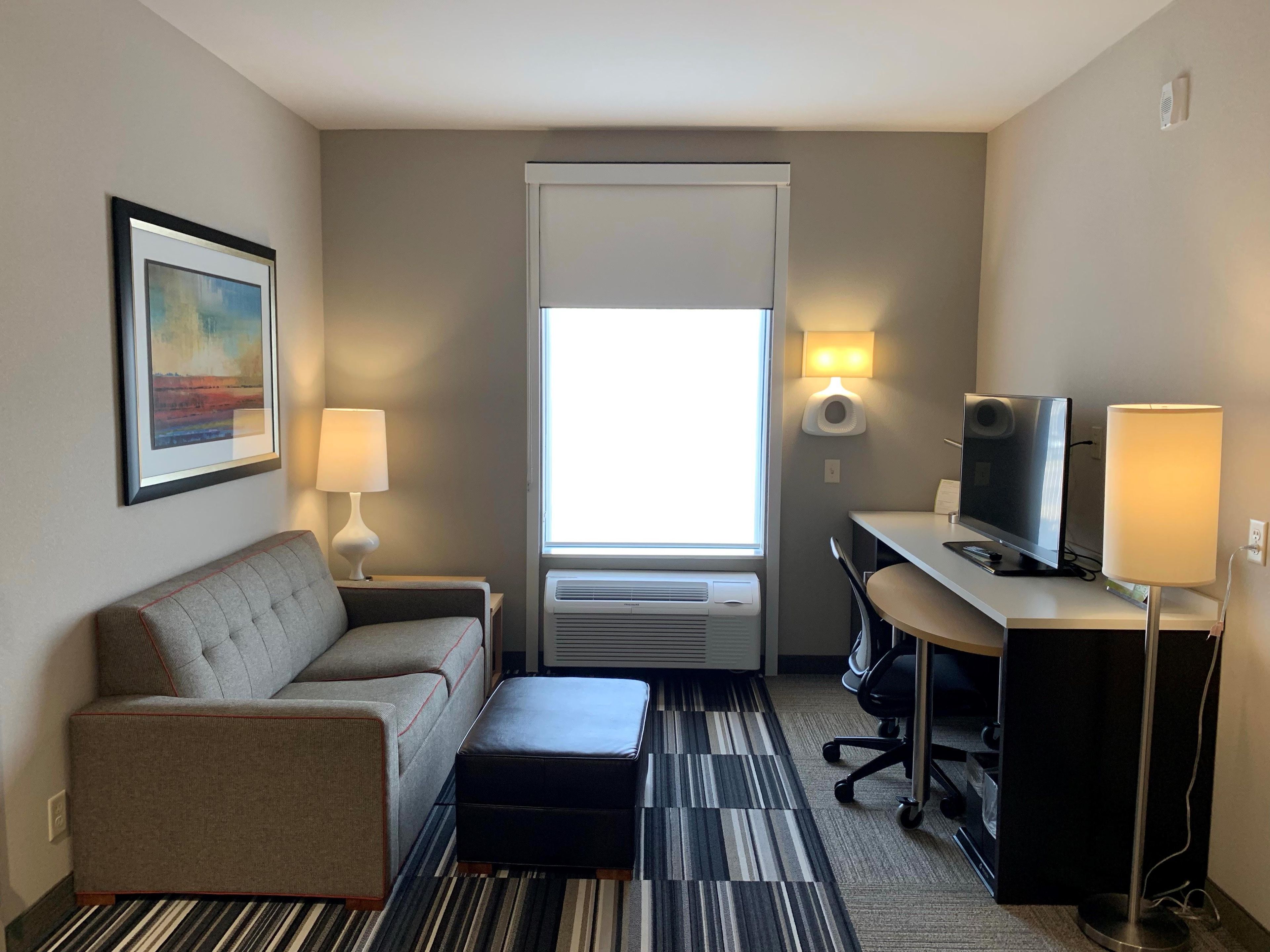 A hotel room with a sofa, a desk with a TV, a lamp, a picture on the wall, and a window with blinds.