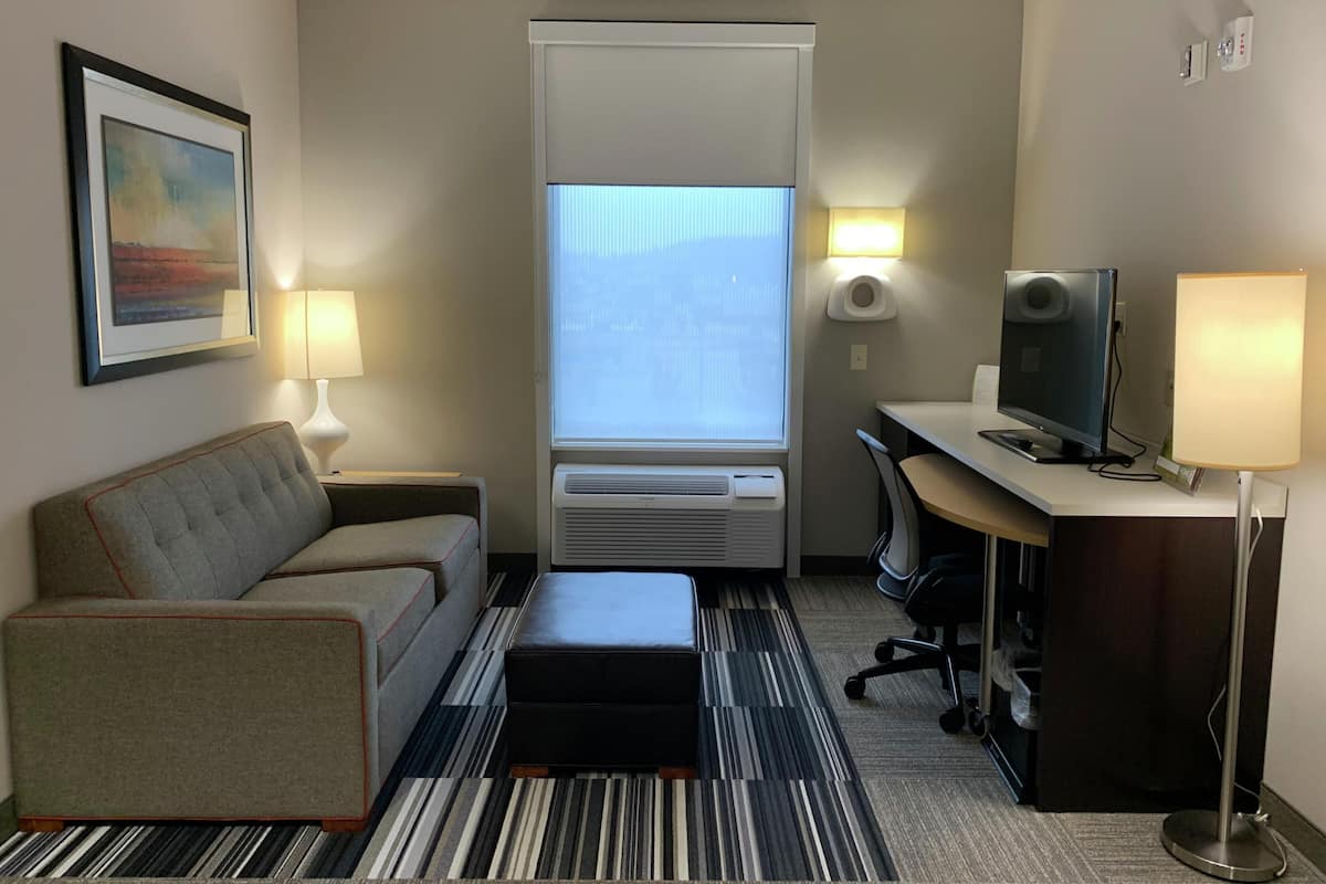 A hotel room with a sofa, a desk with a computer, a television, a window with blinds, an air conditioner, and a lamp.