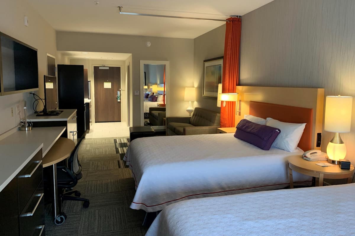 A hotel room with two beds, a desk, a chair, a TV, and a bathroom visible through an open door.