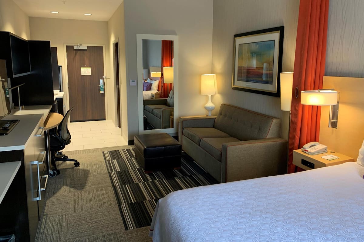 A hotel room with a bed, a desk with a chair, a sofa, a TV, a lamp, and a painting on the wall.