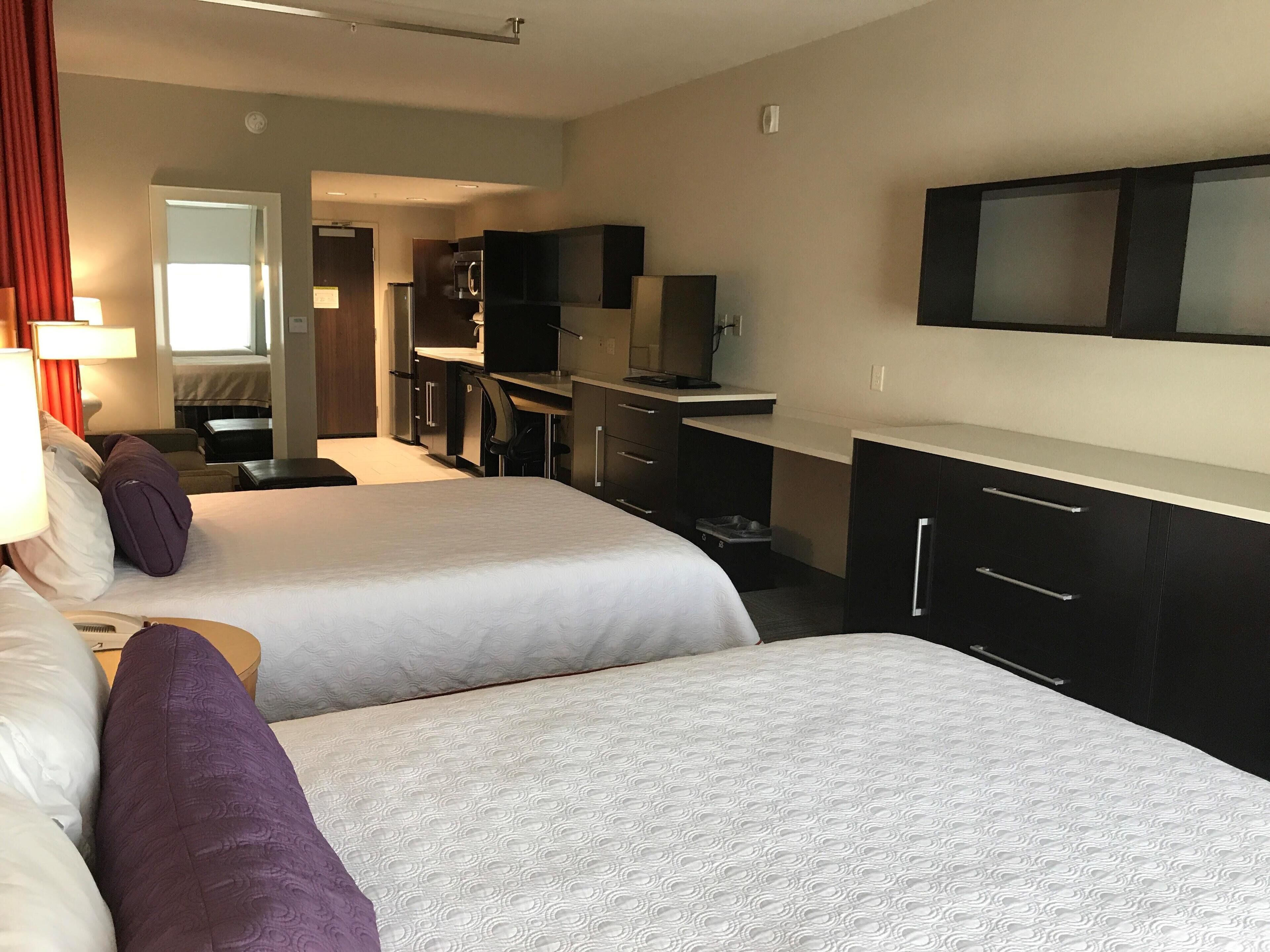 A hotel room with two beds, a desk, a TV, and a kitchenette.