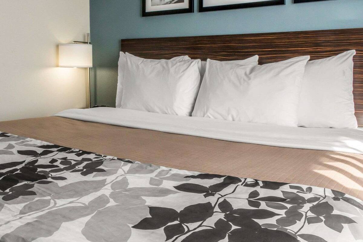 Un lit bien fait, avec du linge de lit blanc et une couverture à motifs floraux, une tête de lit en bois et une lampe fixée au mur.
