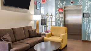A hotel lobby with a brown sofa, a yellow armchair, a flat-screen TV, an exit sign, a motor room sign, and a refrigerator. - Sleep Inn Coney Island (Brooklyn)