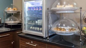 A self-service breakfast station with a refrigerated display case, a double-tiered serving station, and a fruit basket. - Sleep Inn Coney Island (Brooklyn)