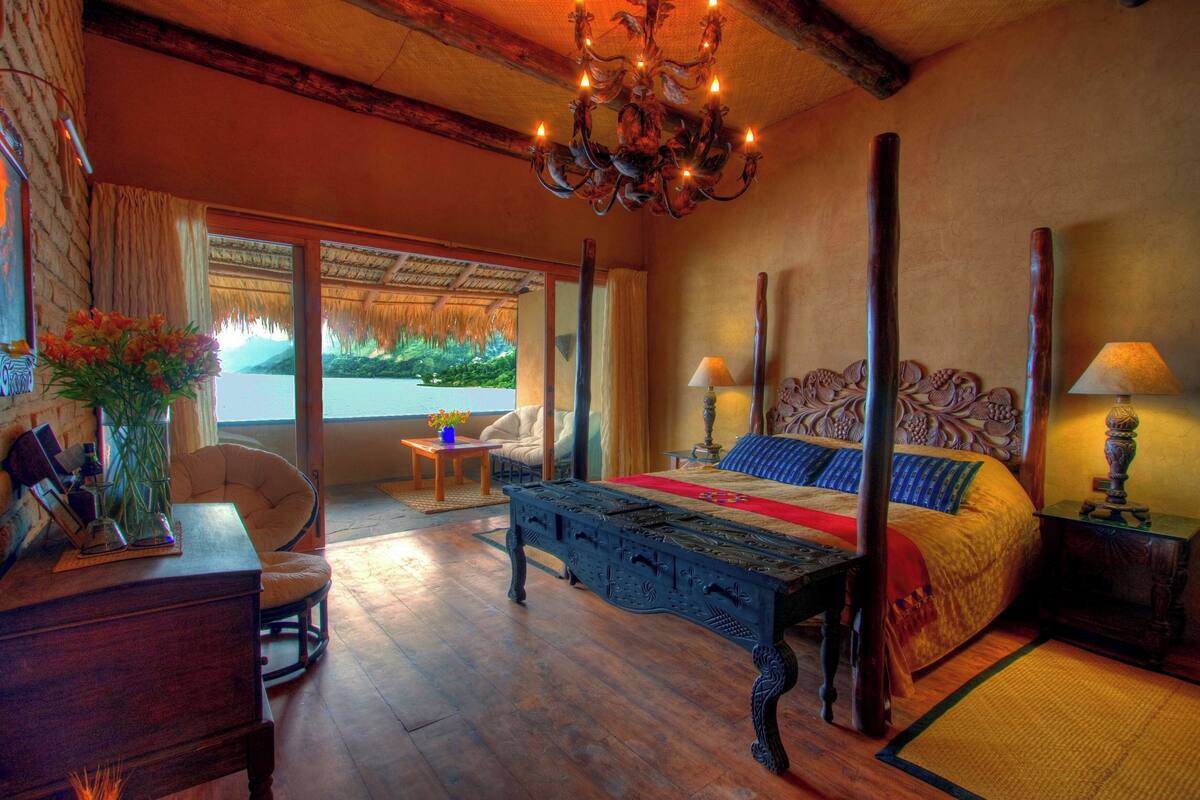 A spacious bedroom with a large bed, a wooden nightstand, a desk, and a chair. There is a chandelier hanging from the ceiling and a window with a view of the outdoors.