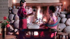 Una habitación acogedora con chimenea de piedra, un banco y una mesa con un jarrón de flores.