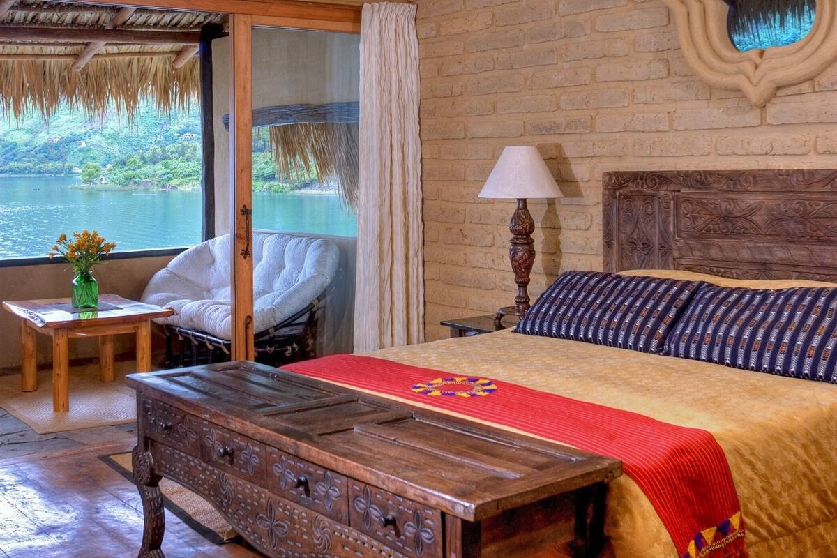 A bedroom with a wooden bed, a nightstand, a chair, a small table with a vase, and a view of a lake through a window.