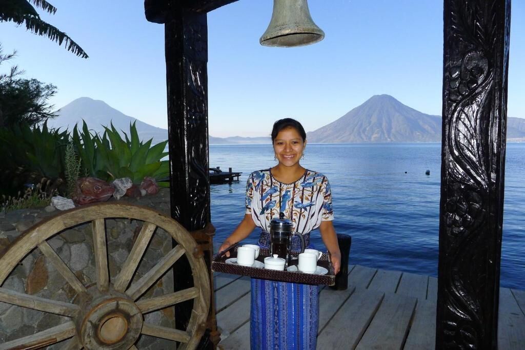 Una persona sirviendo café en una terraza de madera con vista a una montaña y al agua.