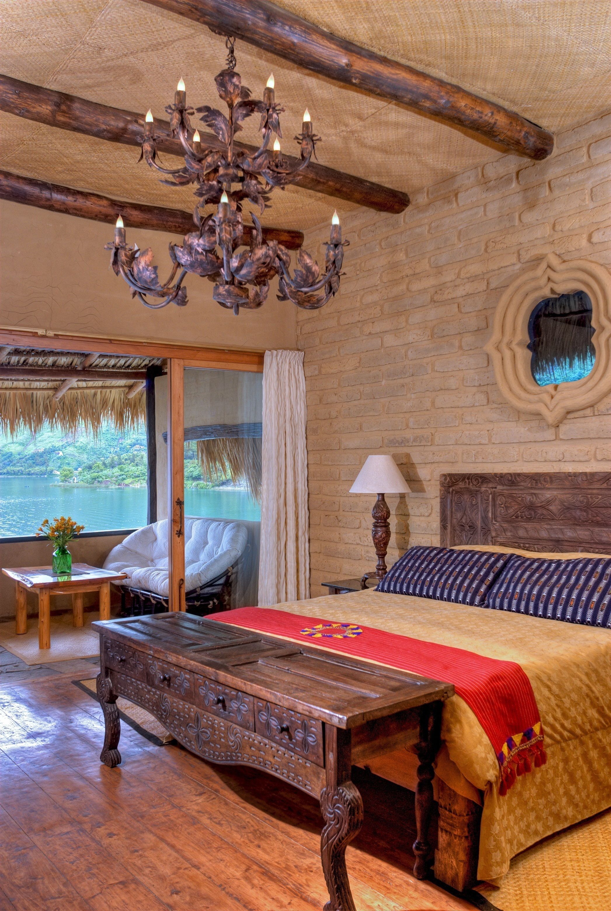 A rustic bedroom with a wooden bed, a chandelier, a wooden nightstand, and a view of a lake through a glass door.