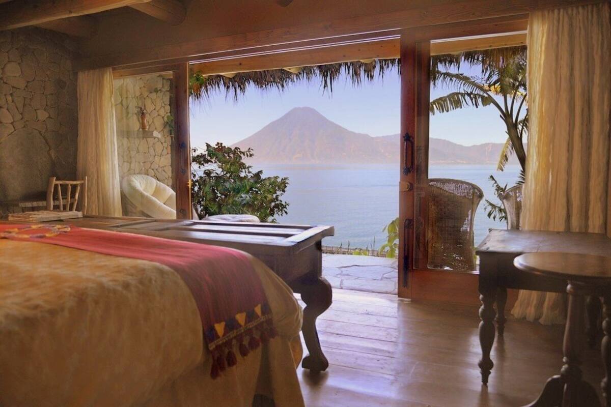 A bedroom with a large bed, a wooden table, and a view of a body of water and a mountain.