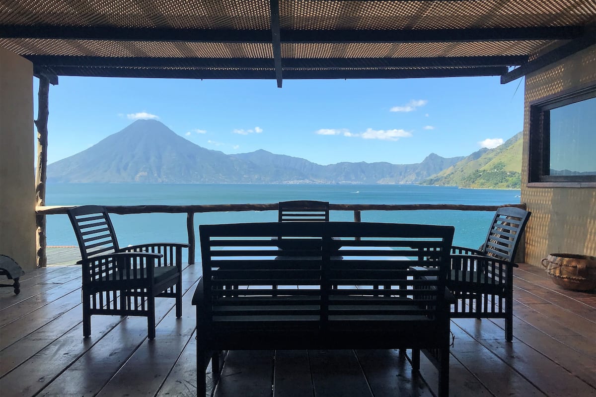 A wooden deck with a table and chairs, overlooking a calm sea and a mountain range.