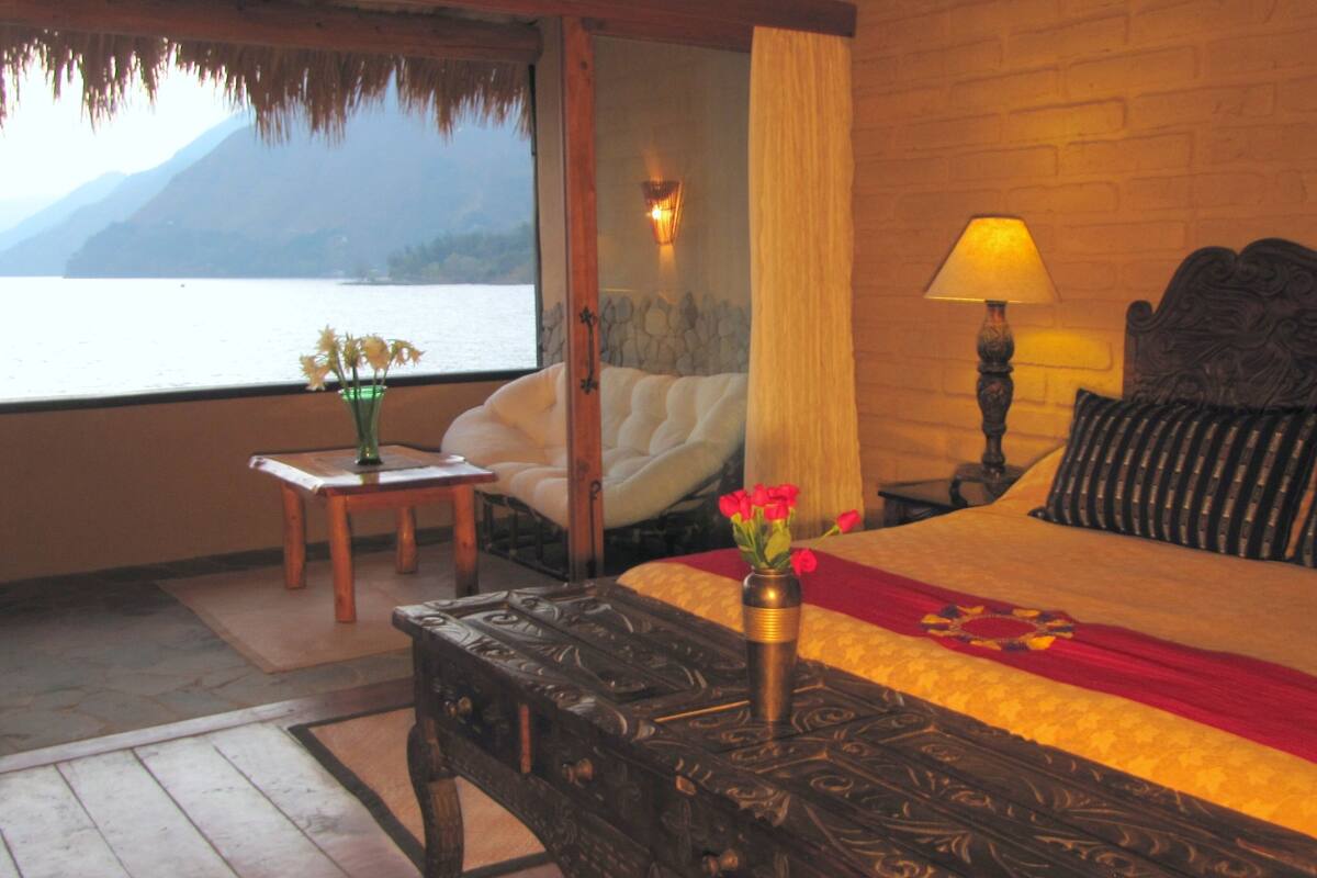 A bedroom with a bed, a nightstand, a lamp, a vase with flowers, a view of the sea, and a wooden ceiling.
