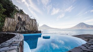 Una piscina infinita de color azul cristalino, con borde de piedra, una estructura de techo de paja y una montaña al fondo.