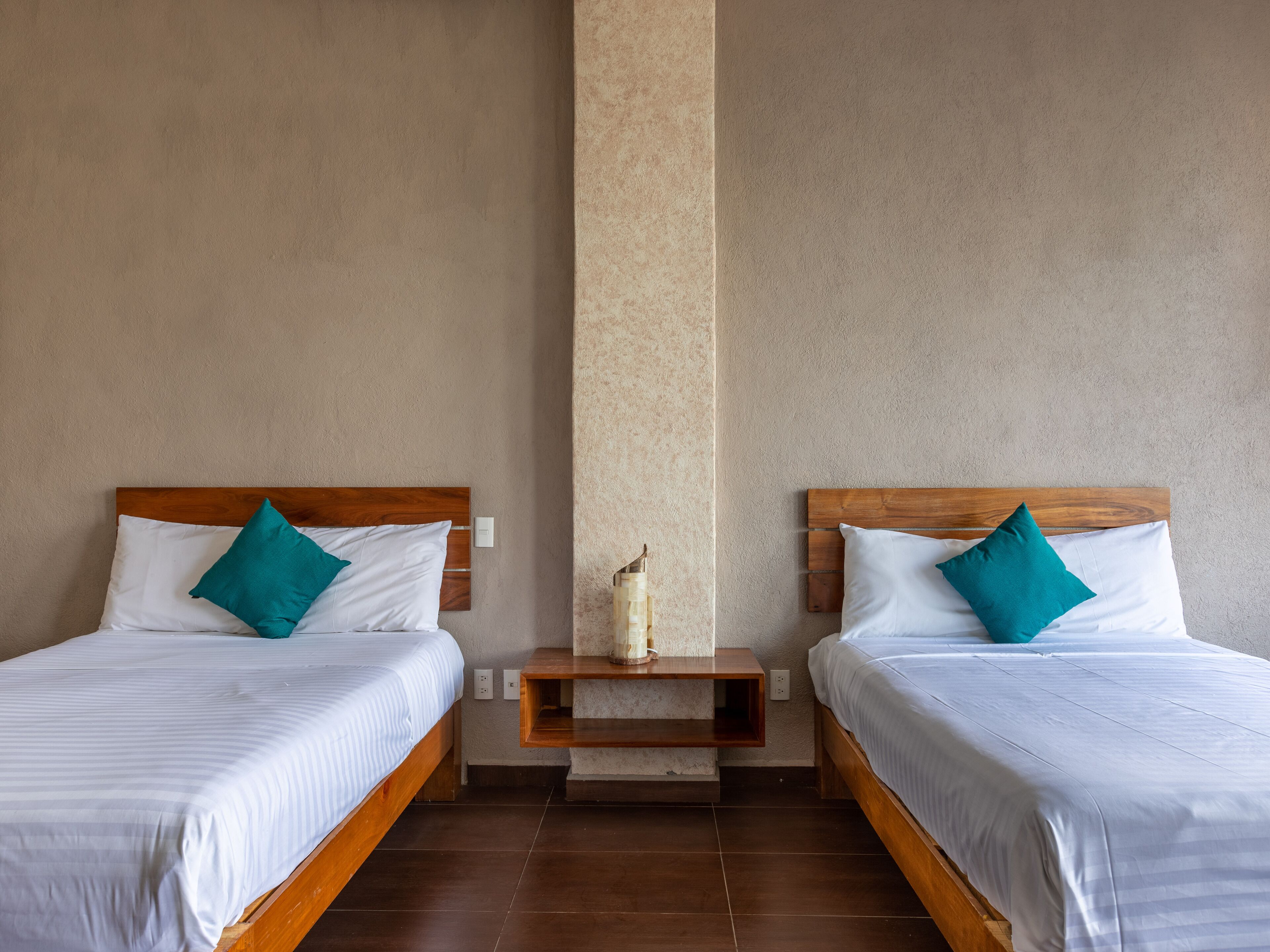 Habitación de hotel con dos camas individuales, cada una con ropa blanca y almohadas color turquesa, un cabecero de madera y una mesita de noche con lámpara.