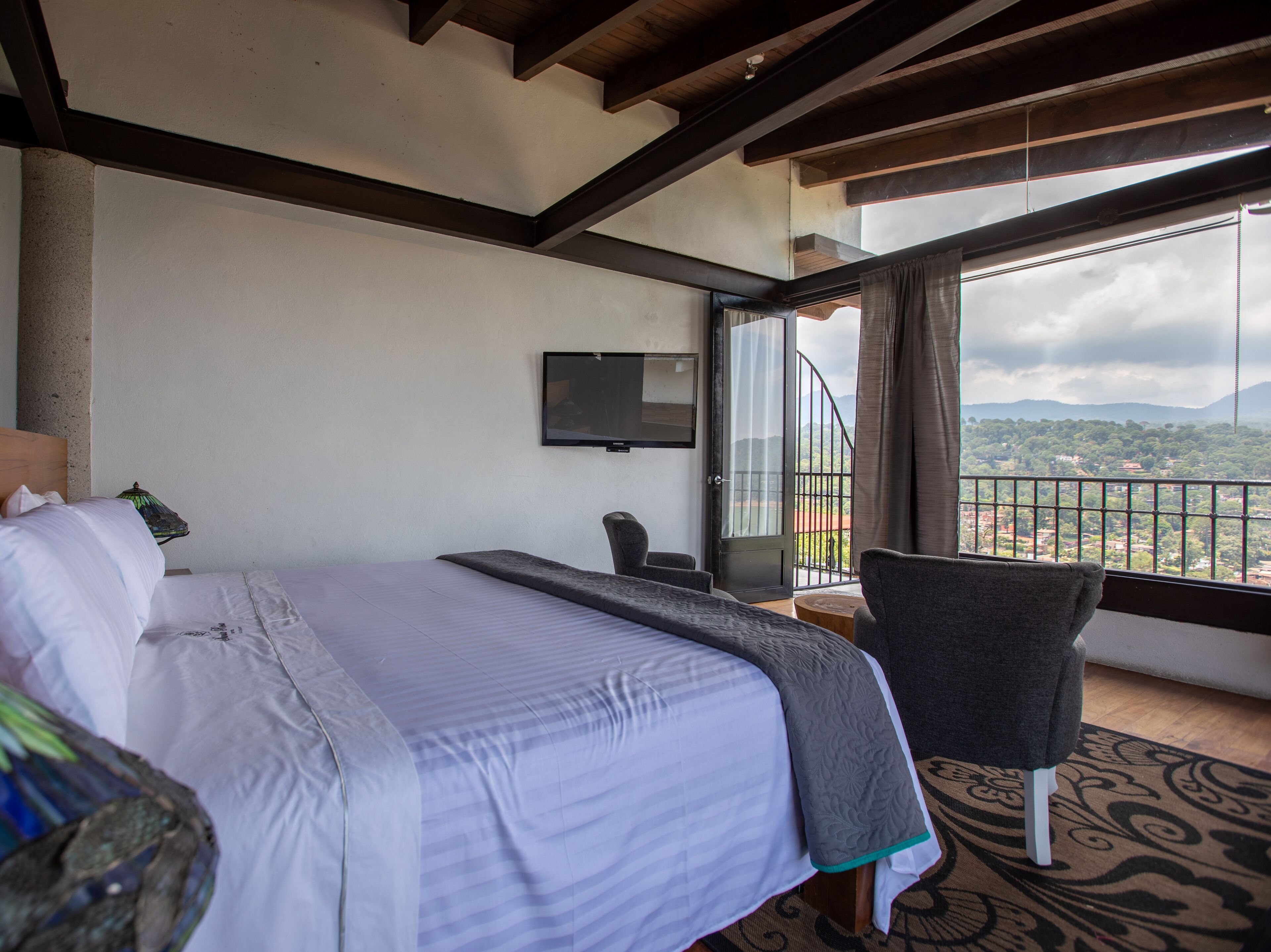 A hotel room with a bed, a TV, a chair, and a balcony with a view of the landscape.