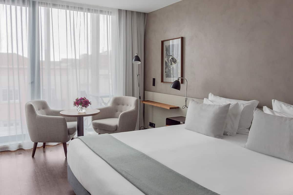 Een moderne hotelkamer met een groot bed, twee fauteuils, een klein rond tafeltje met een bloevas, een bureau met een lamp en een groot raam met dunne gordijnen.