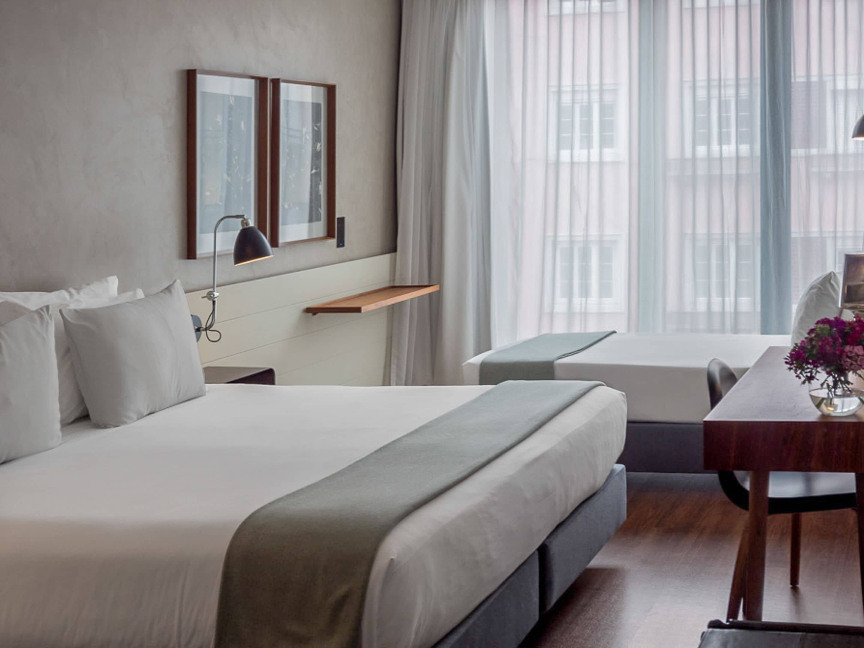 A modern hotel room with a large bed, a desk, and a chair. There is a window with sheer curtains and a view of buildings outside.