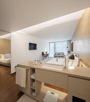 Una habitación de hotel moderna con una cama grande, un área de estar separada y un baño con un tocador amplio y lavamanos.