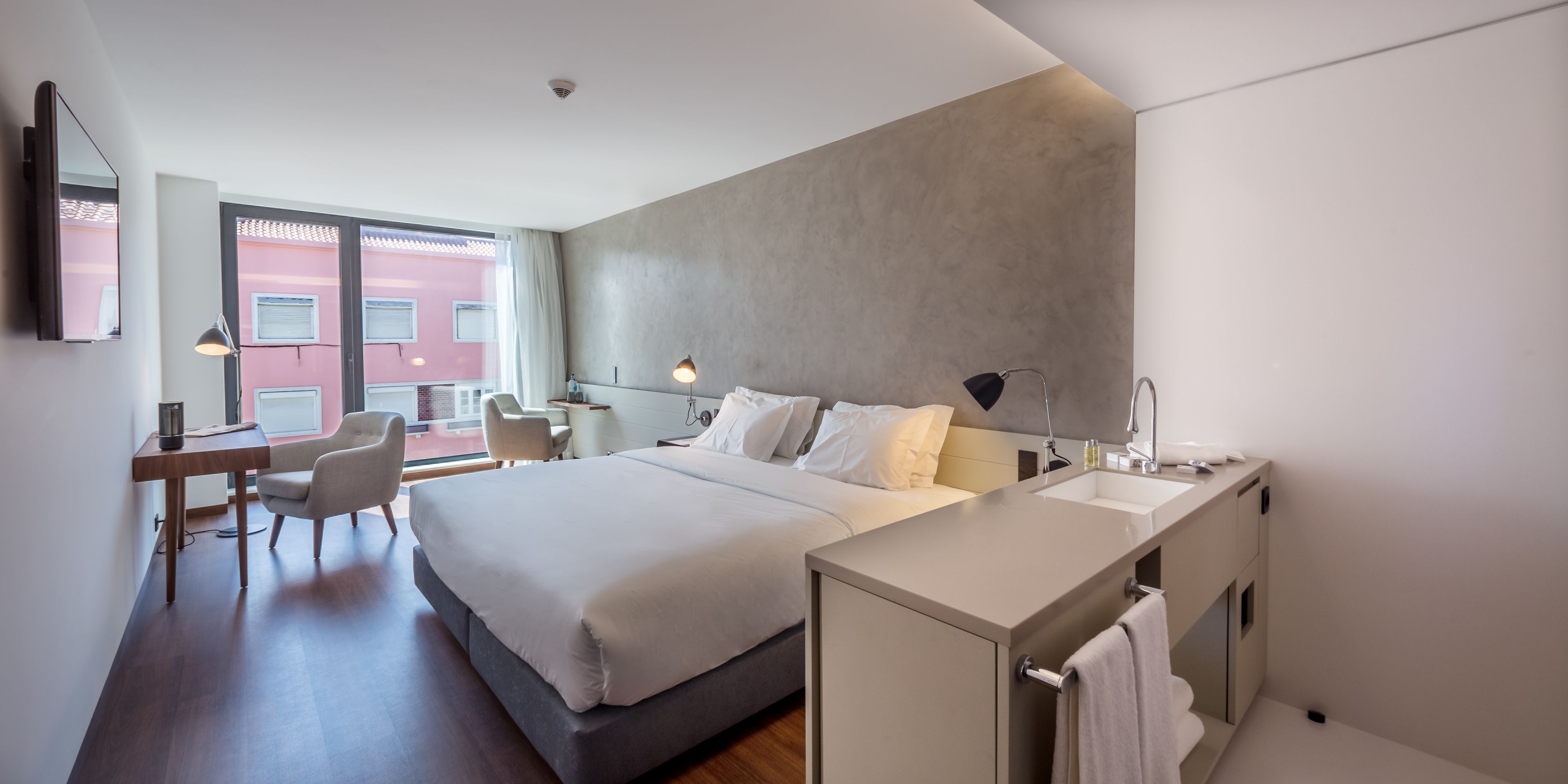 A modern hotel room with a large bed, bedside table, and a small desk with a chair.
