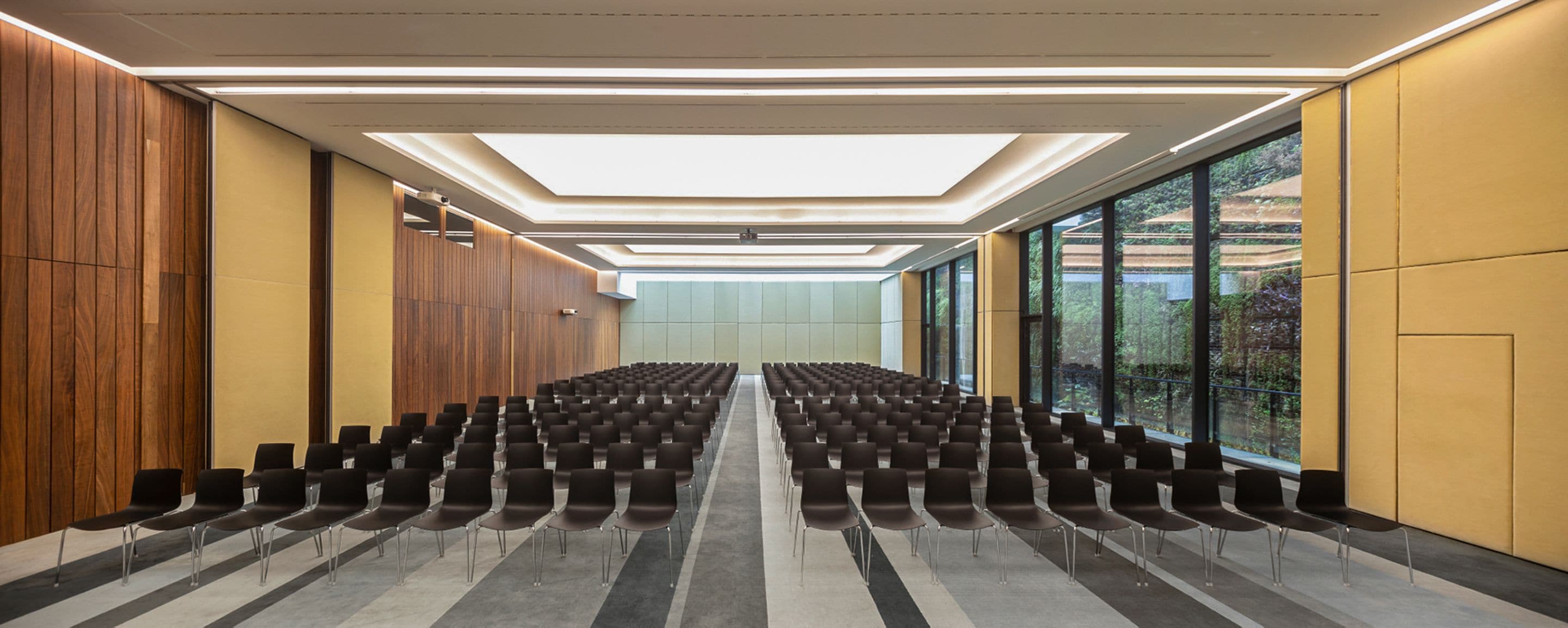 A conference room with rows of chairs, a skylight ceiling, and large windows offering a view of the outdoors.
