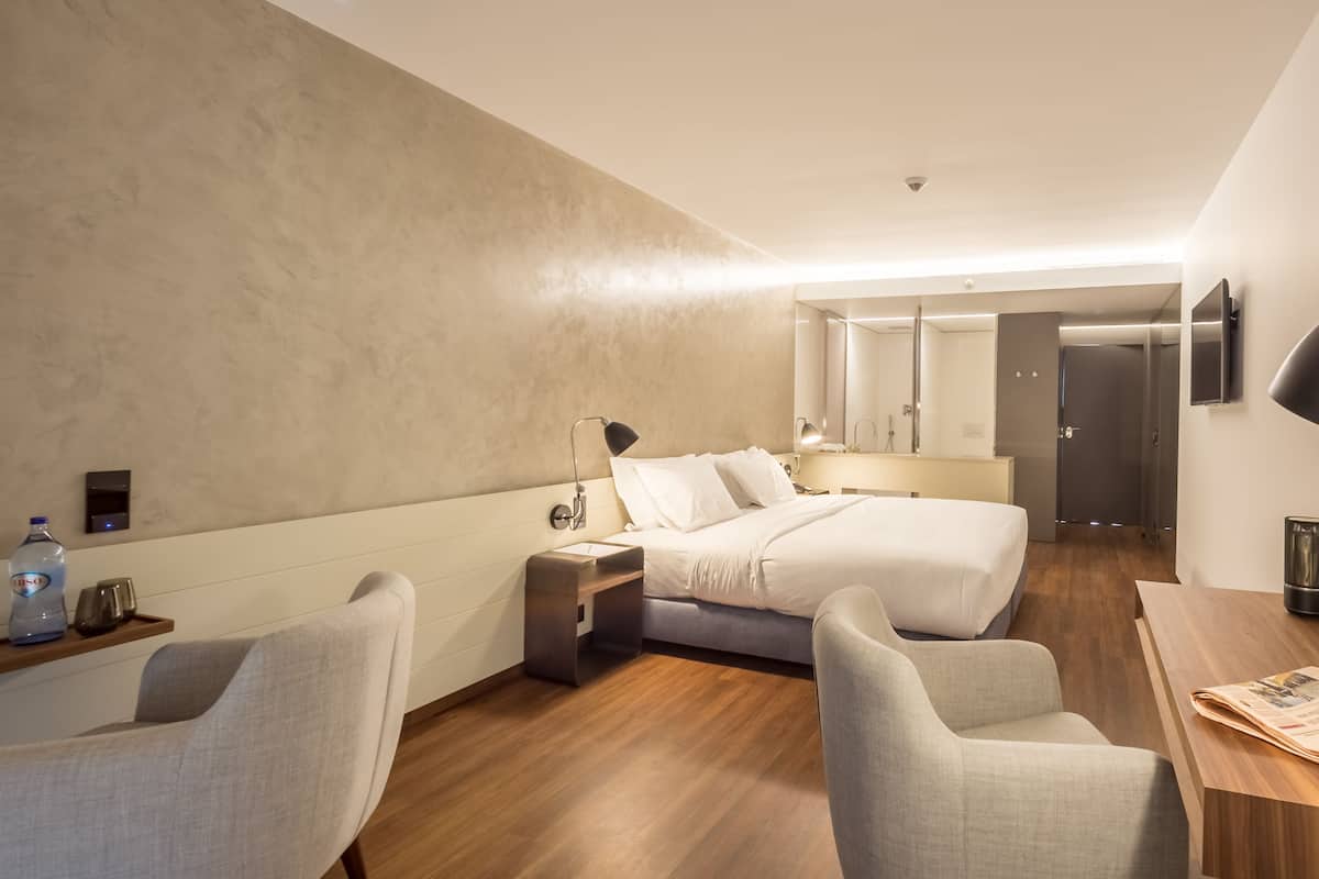 Een moderne hotelkamer met een bed, twee stoelen, een bureau met een krant, een nachtkastje met een fles en een wandlamp.