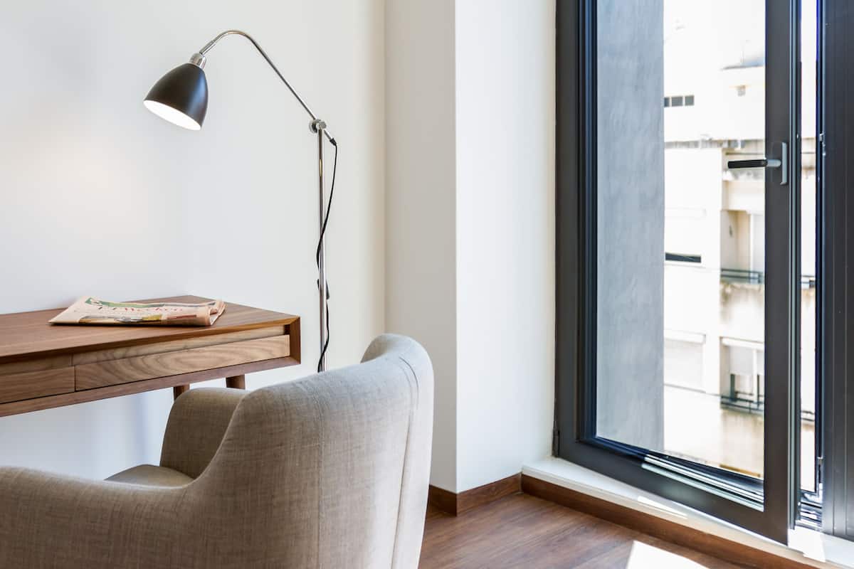 Een moderne kamer met een houten bureau, een zwarte lamp, een beige stoel en een groot raam met uitzicht op gebouwen.