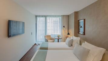 Ein Hotelzimmer mit zwei Betten, einem an der Wand befestigten Fernseher, einem Sessel, einem kleinen Tisch mit Lampe und einem Fenster mit Vorhängen.