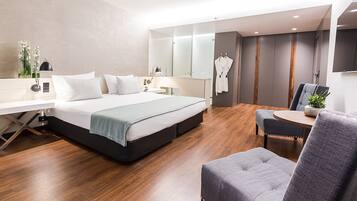 A modern hotel room with a bed, bedside table, chair, and a small table with a plant.
