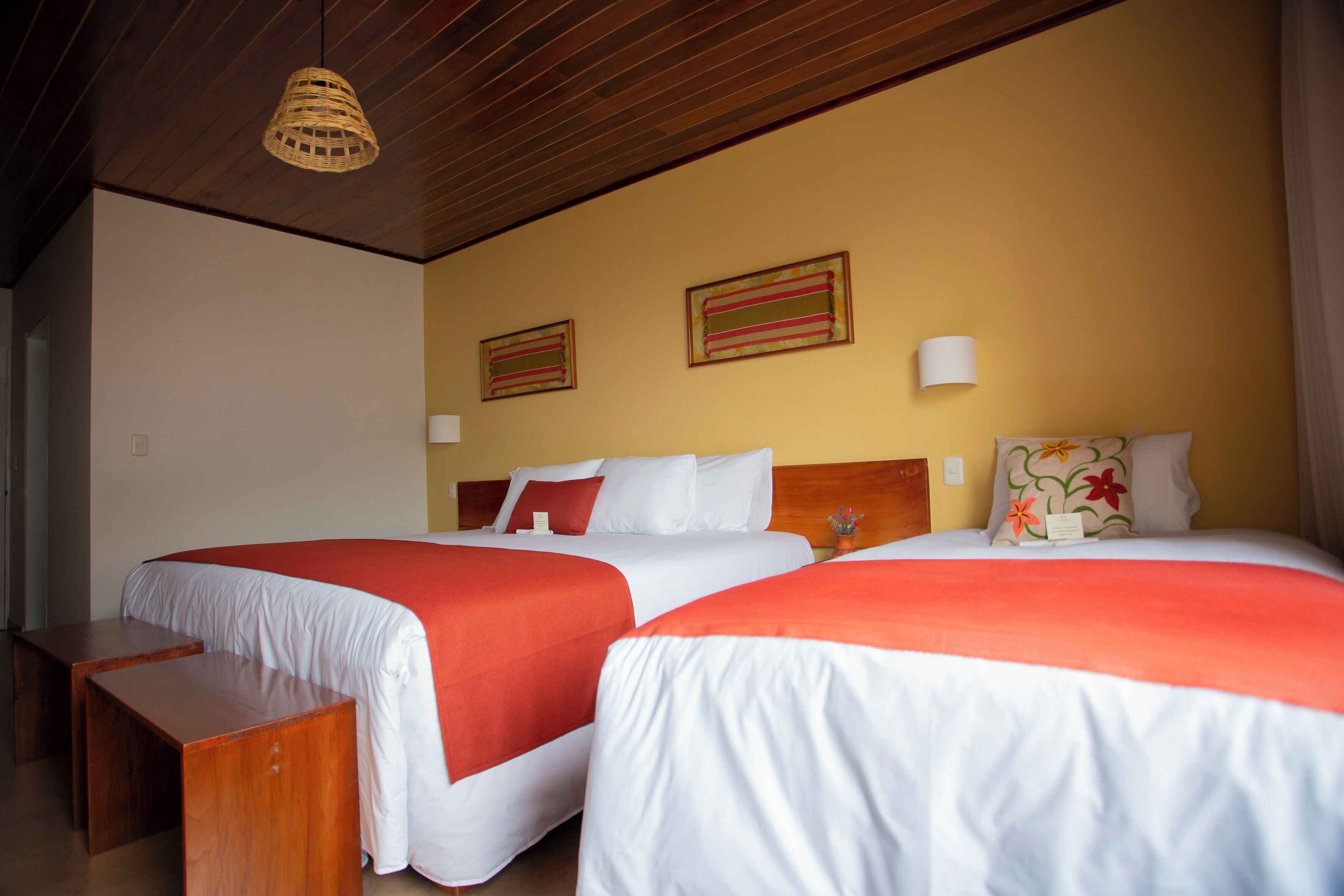 A hotel room with two beds, wooden headboards, a woven ceiling light, and two framed artworks on the wall.
