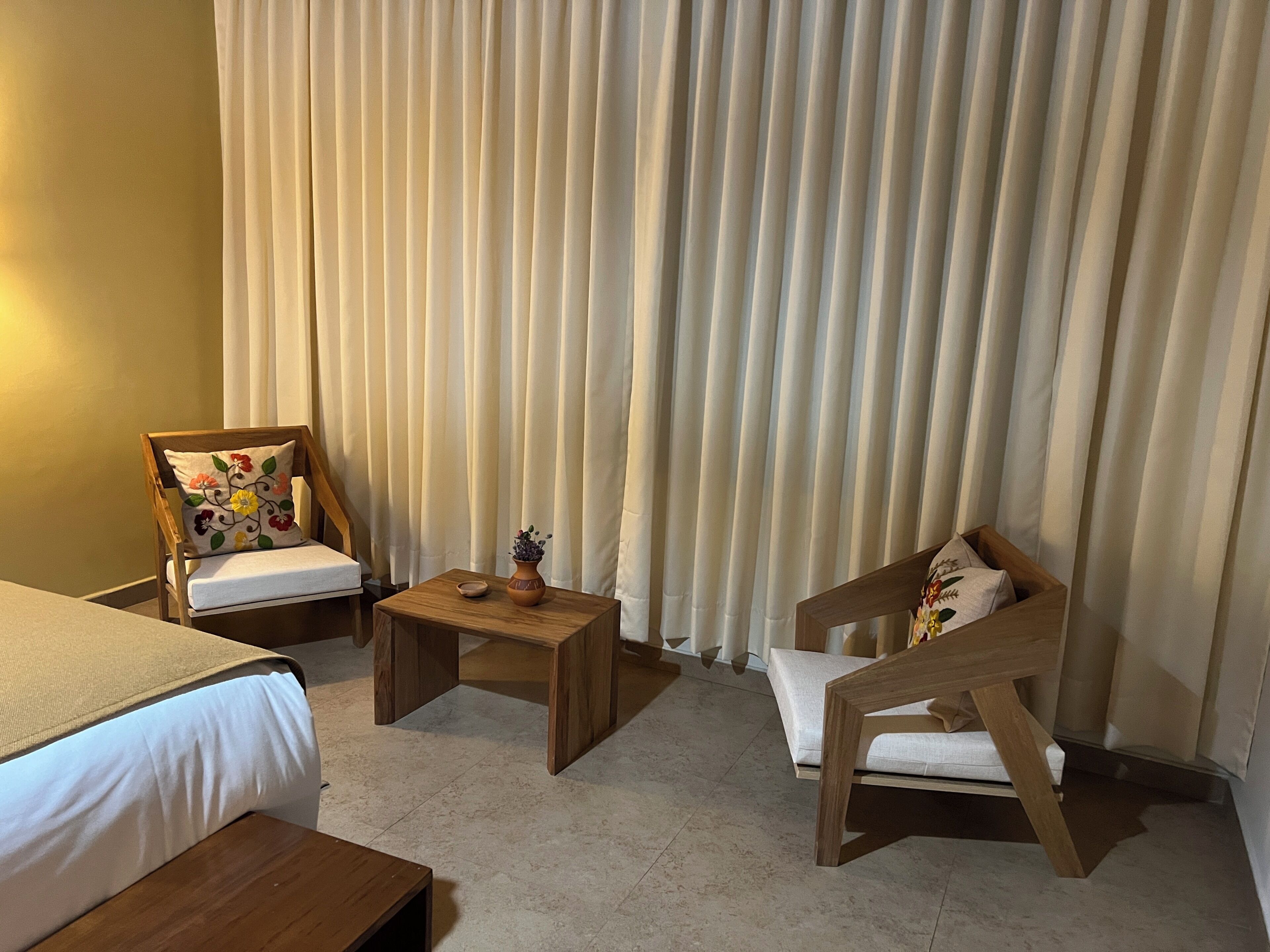 Habitación de hotel con cama, dos sillas de madera, una mesita pequeña y un ventanal con cortinas.