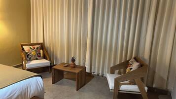 Habitación de hotel con cama, dos sillas de madera, una mesita pequeña y un ventanal con cortinas.