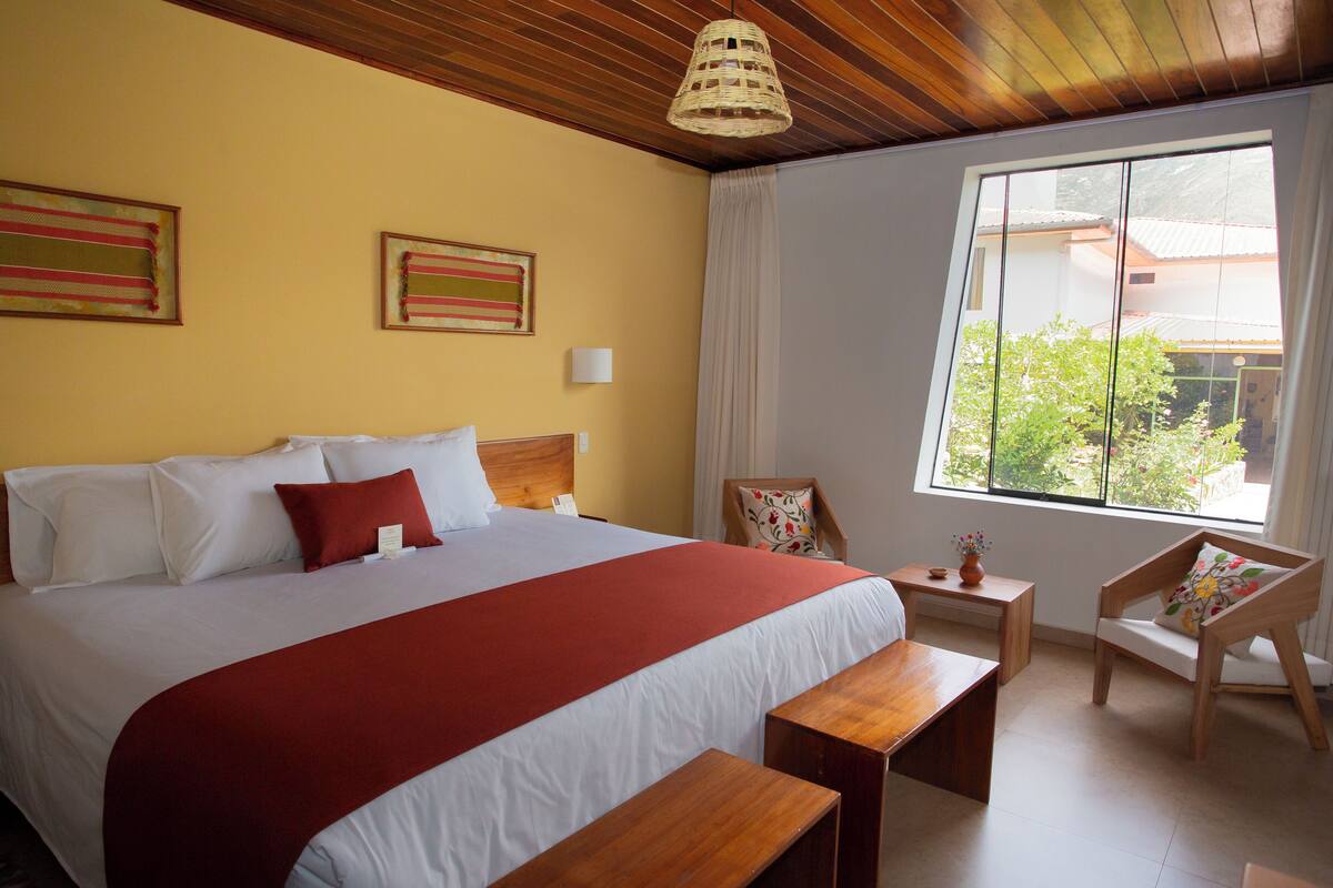 Une chambre d’hôtel avec un grand lit, une tête de lit en bois, une fenêtre donnant sur un beau paysage, une petite table et une chaise.