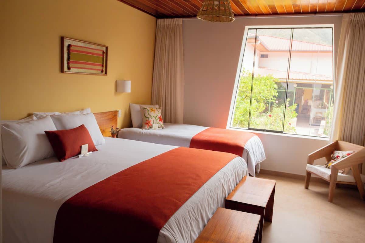 Une chambre d’hôtel avec deux lits, un banc en bois, une chaise, une fenêtre donnant sur un paysage verdoyant et une lampe fixée au mur.