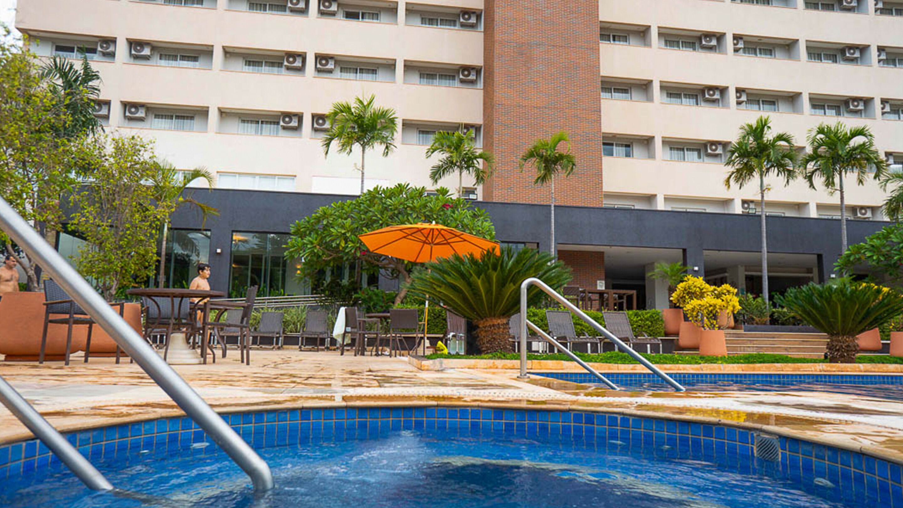 A swimming pool with lounge chairs and an umbrella, surrounded by palm trees and a building with multiple floors and air conditioning units.