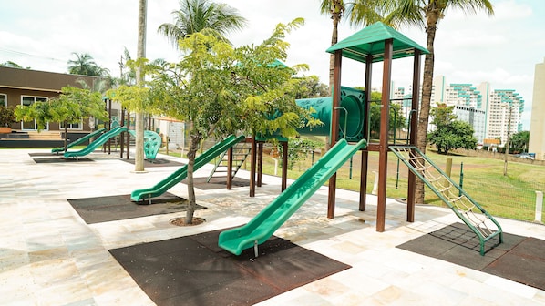 A playground with multiple slides and climbing structures, surrounded by palm trees and a paved area.