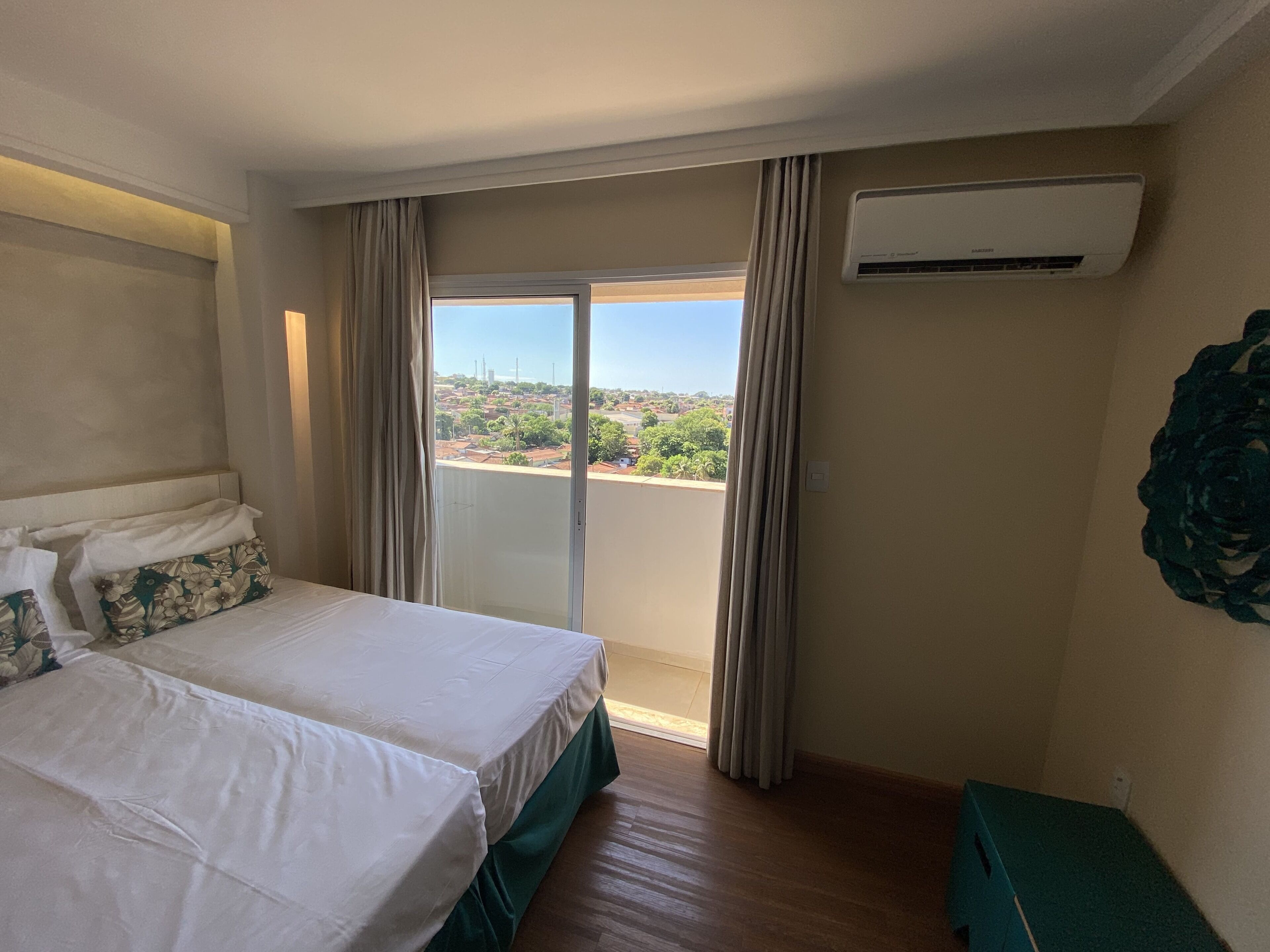 Ein Hotelzimmer mit Bett, Blick auf eine Stadtkulisse durch ein großes Fenster und einer Klimaanlage an der Wand.