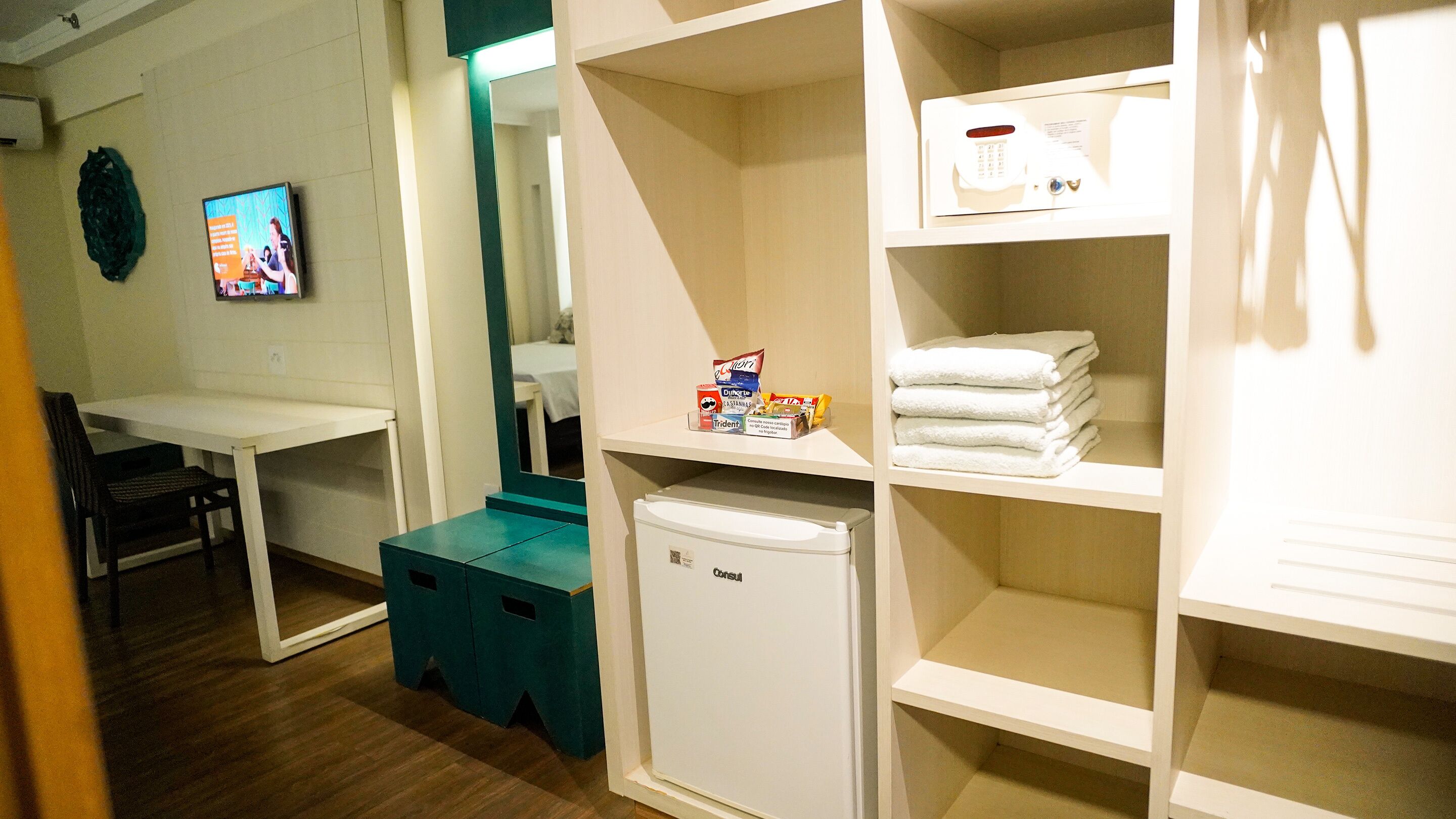 A hotel room with a desk, a small fridge, and shelves with towels and toiletries.