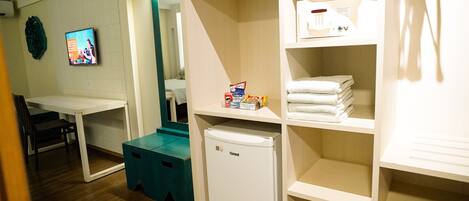 A hotel room with a desk, a small fridge, and shelves with towels and toiletries.