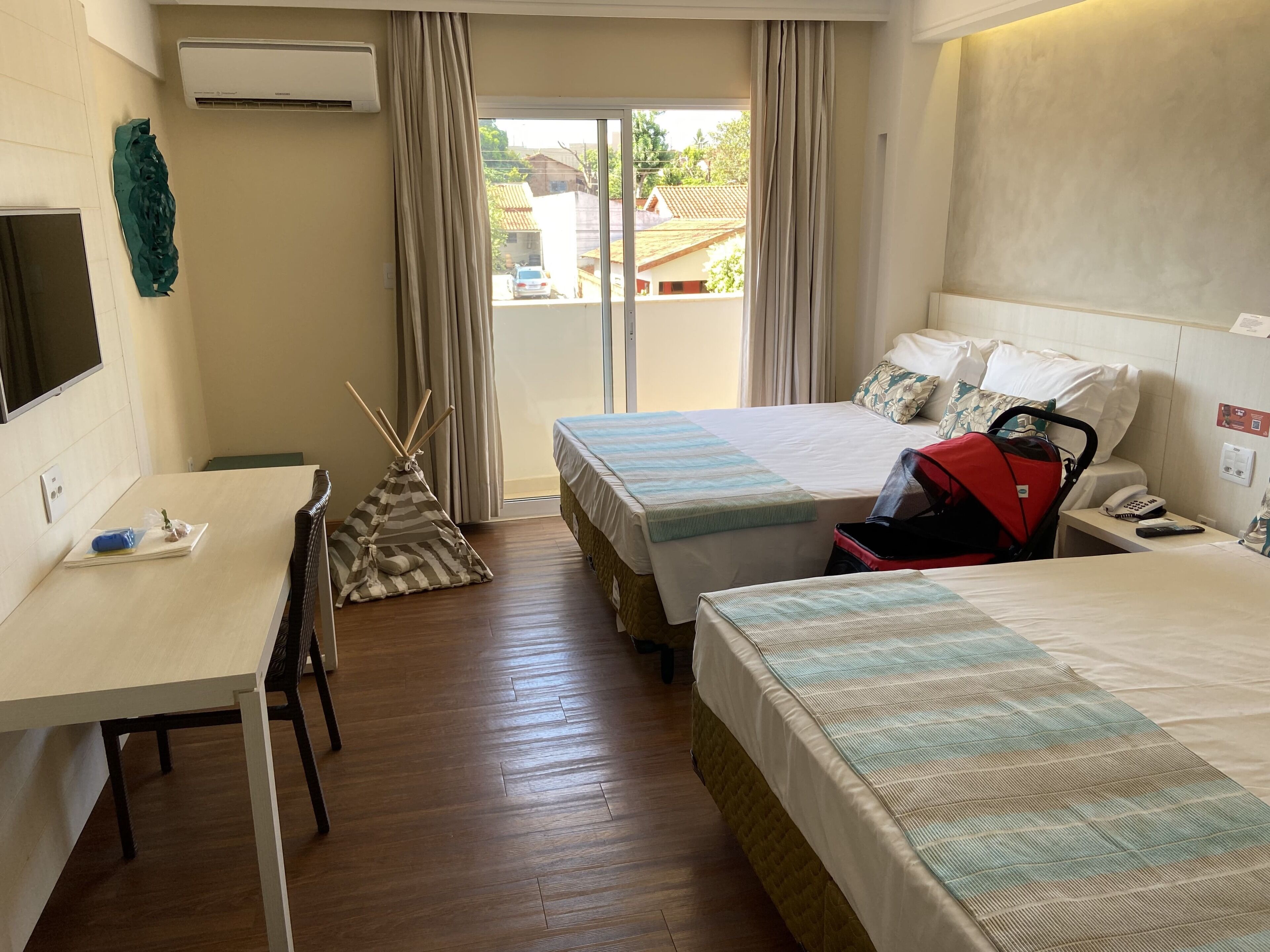 A hotel room with two beds, a desk, a chair, a red suitcase, and a view of the outside.