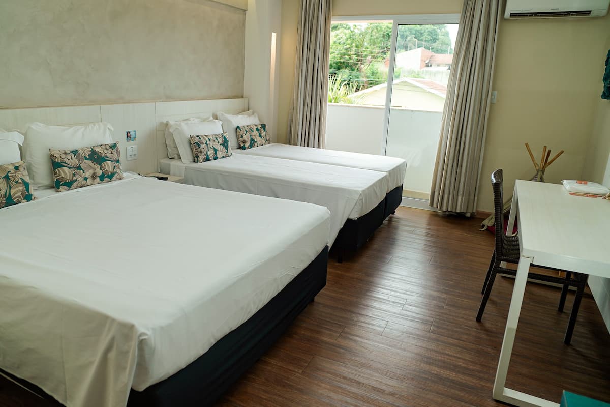 A hotel room with three single beds, a wooden floor, a white table, and a view of the outside.