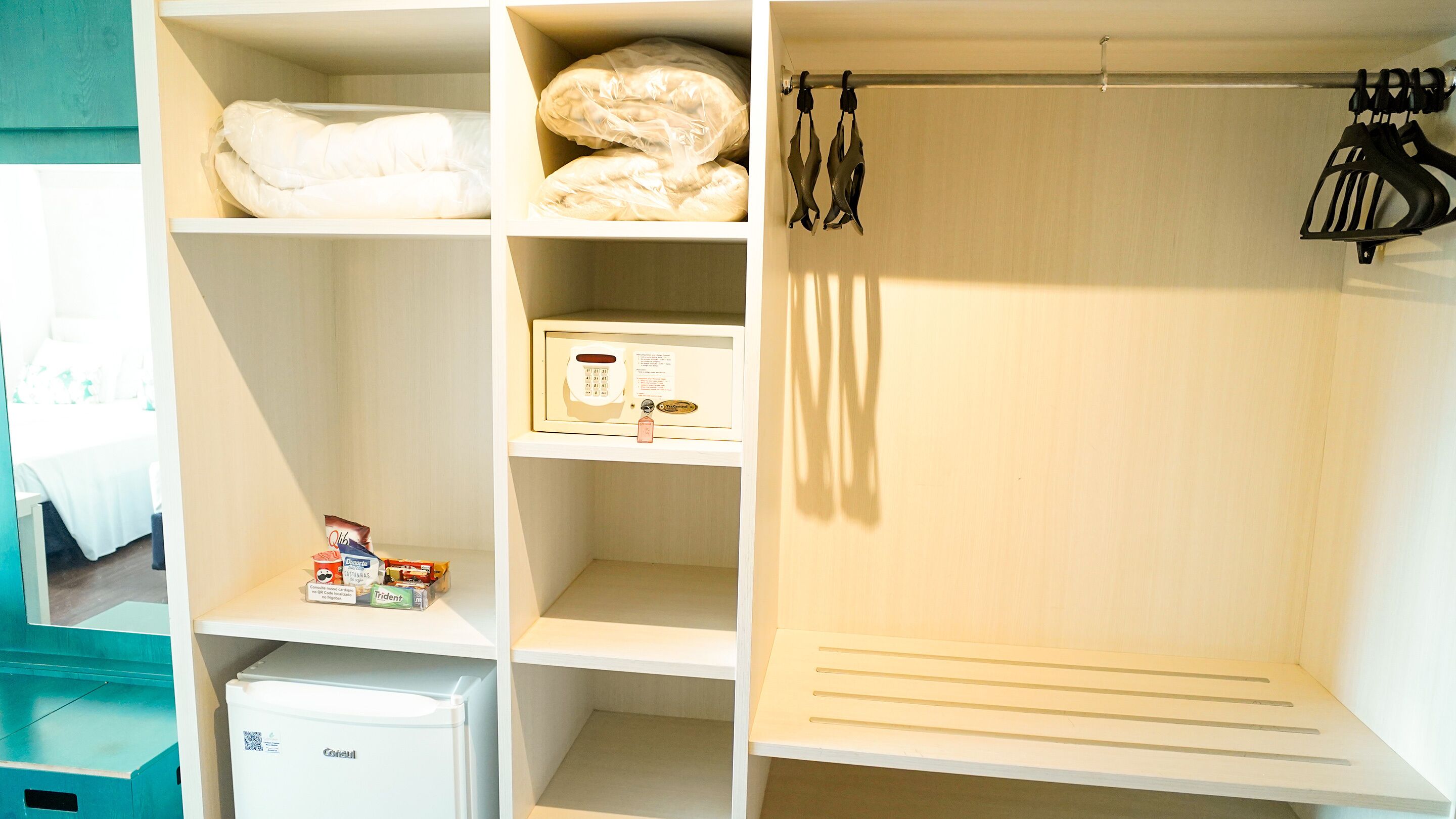 A hotel room with a bed, a small refrigerator, shelves with toiletries, and a shower area with a towel rack.